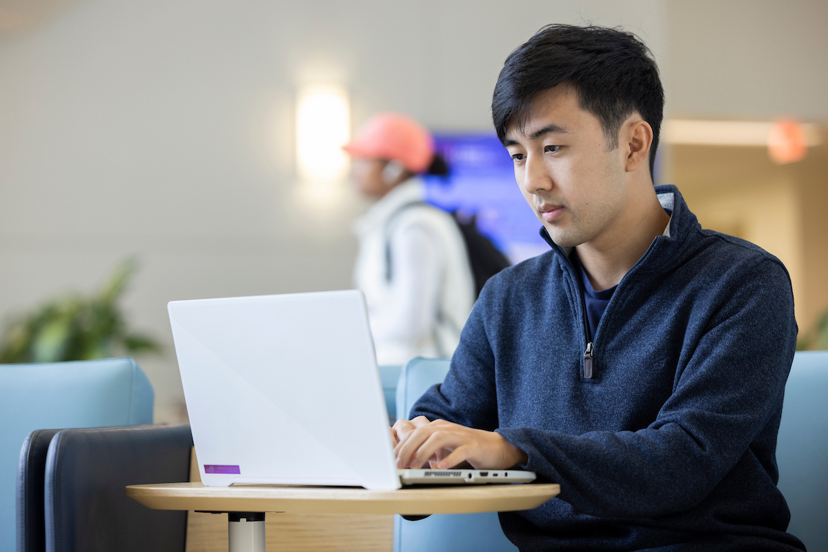 A person seated at a table using a laptop in what appears to be a lounge or waiting area. Another individual is blurred in the background, walking past, wearing a cap and backpack.