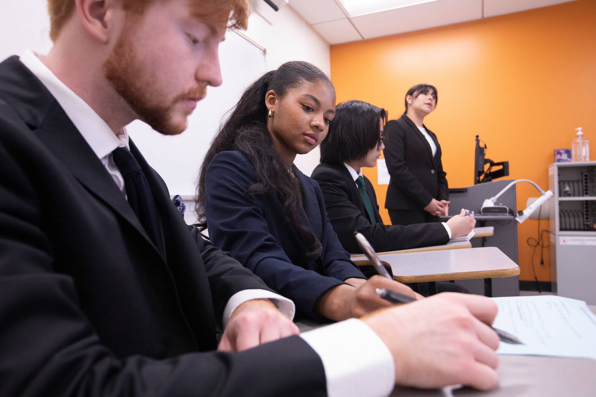Three students in business attire take notes while a person in the far background speaks.