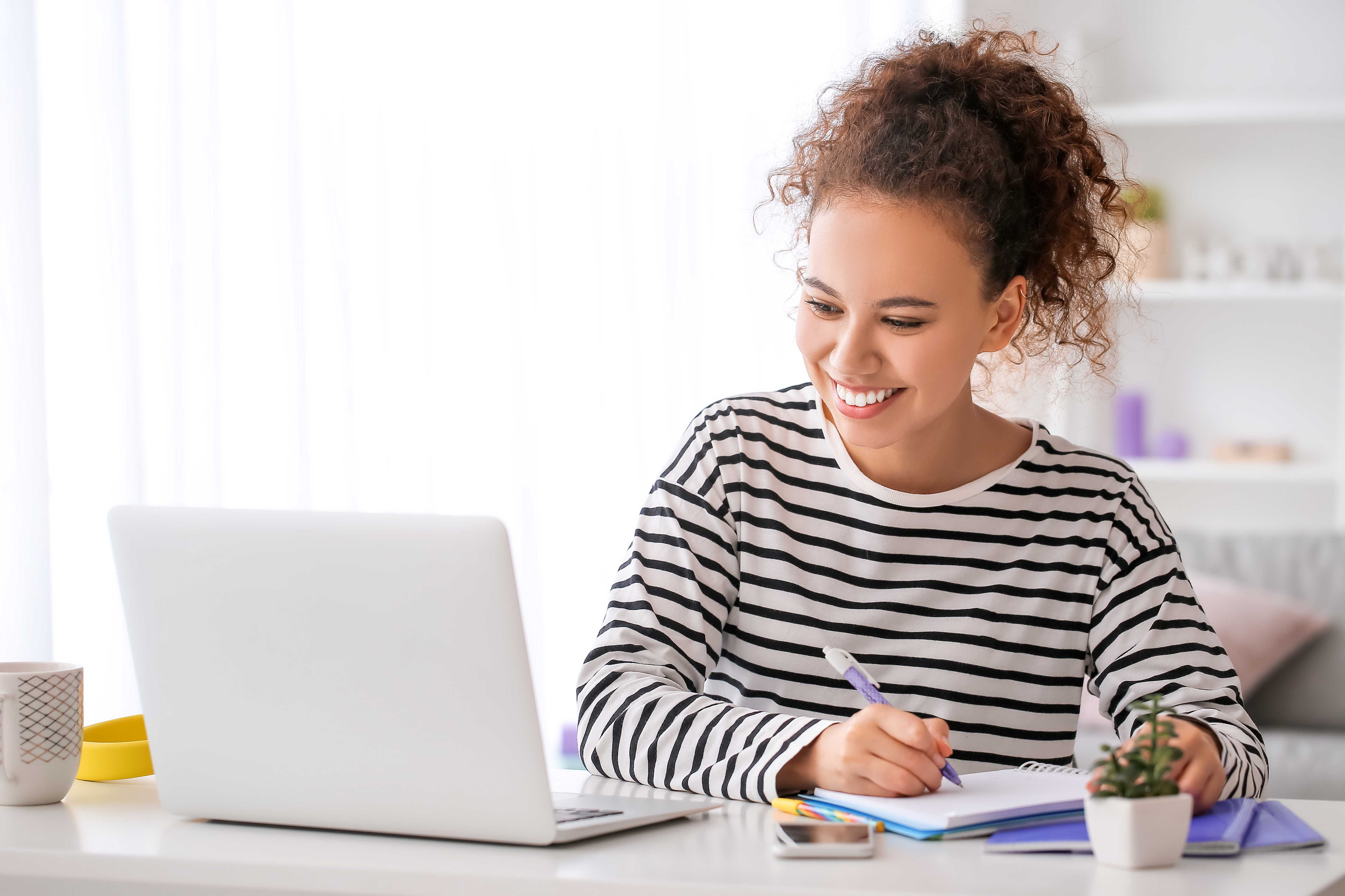 A woman smiles at her laptop, taking notes on an online course.