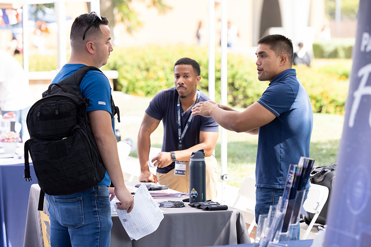 Three men discussing at an outdoor informational booth on a college campus