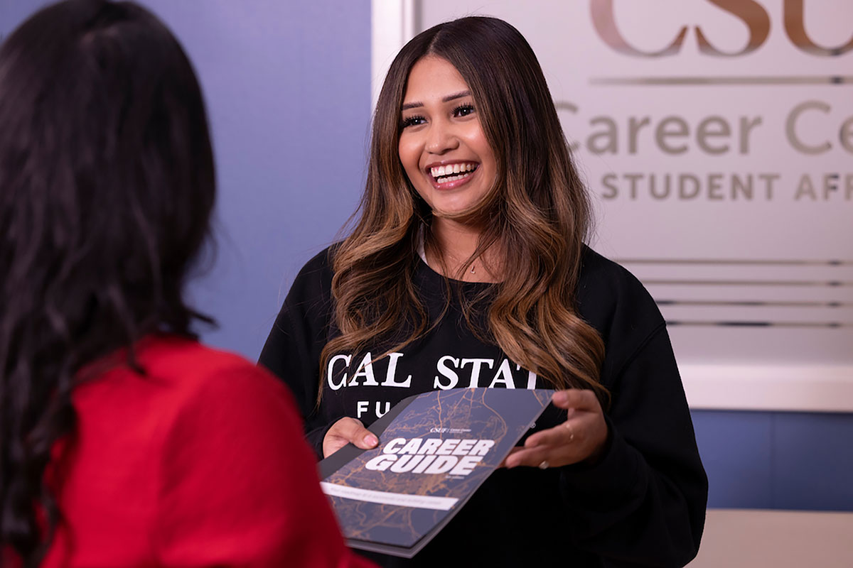 Smiling student holding a 'Career Guide' booklet while talking to an advisor at the CSUF Career Center.